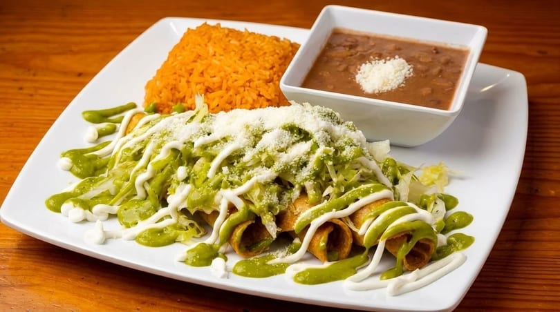 Crispy taquitos topped with green sauce, crema, lettuce, and cheese, served with rice and beans.