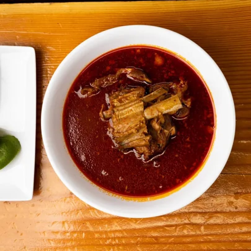 A white bowl of rich red birria stew with chunks of meat, next to a small dish with lime wedges.